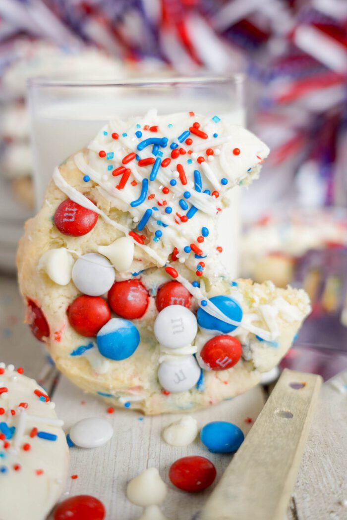 A cookie decorated with red, white, and blue candy pieces and sprinkles, partially eaten, next to a glass of milk.