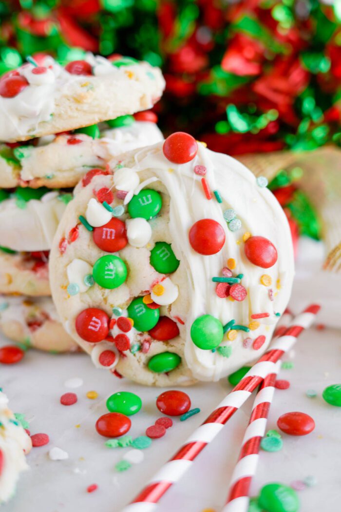 A stack of festive cookies covered in white chocolate and decorated with red, green, and white M&M's, sprinkles, and candies. Red and white striped straws are placed beside the cookies.