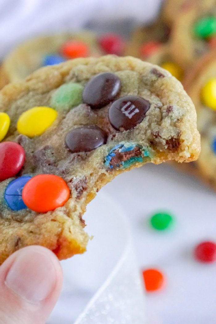 A hand holds a partially eaten chocolate chip cookie with colorful candy-coated chocolates (M&Ms) embedded in it. Additional candies are scattered in the background.