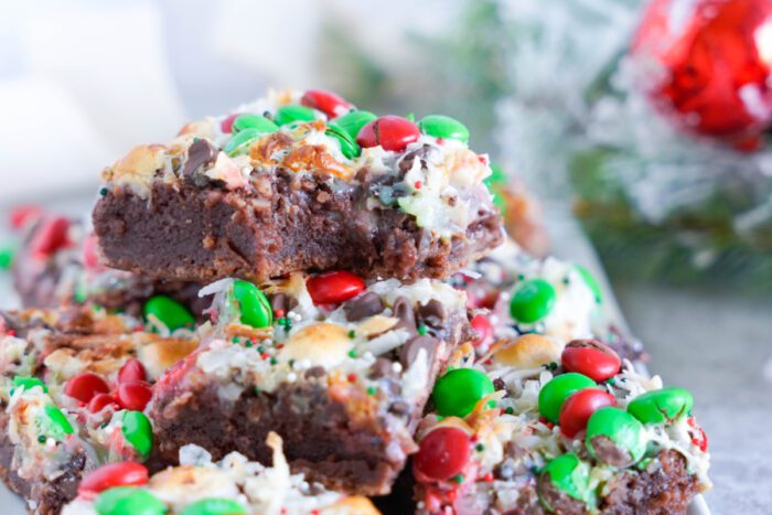 A stack of holiday-themed brownies topped with red and green candies, coconut flakes, and chocolate chips on a white surface with festive decorations in the background.