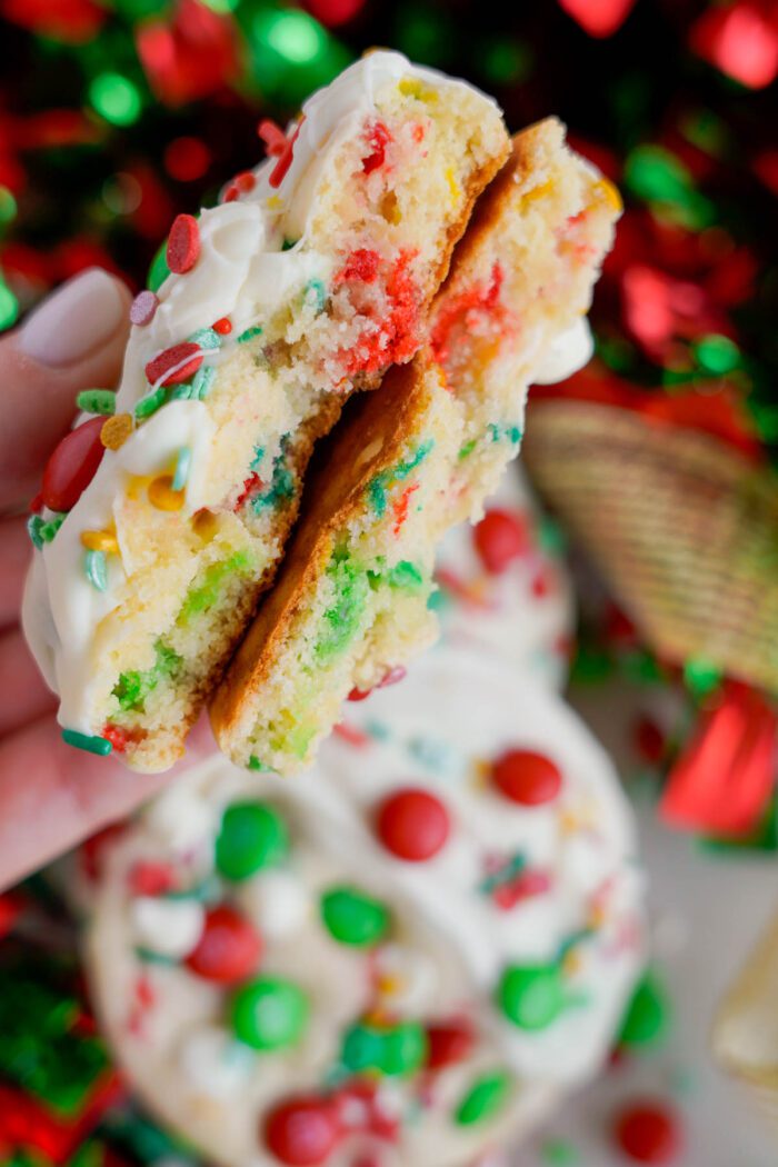 A hand holding a split cookie with colorful sprinkles and white chocolate. Decorated cookies with similar style are blurred in the background.