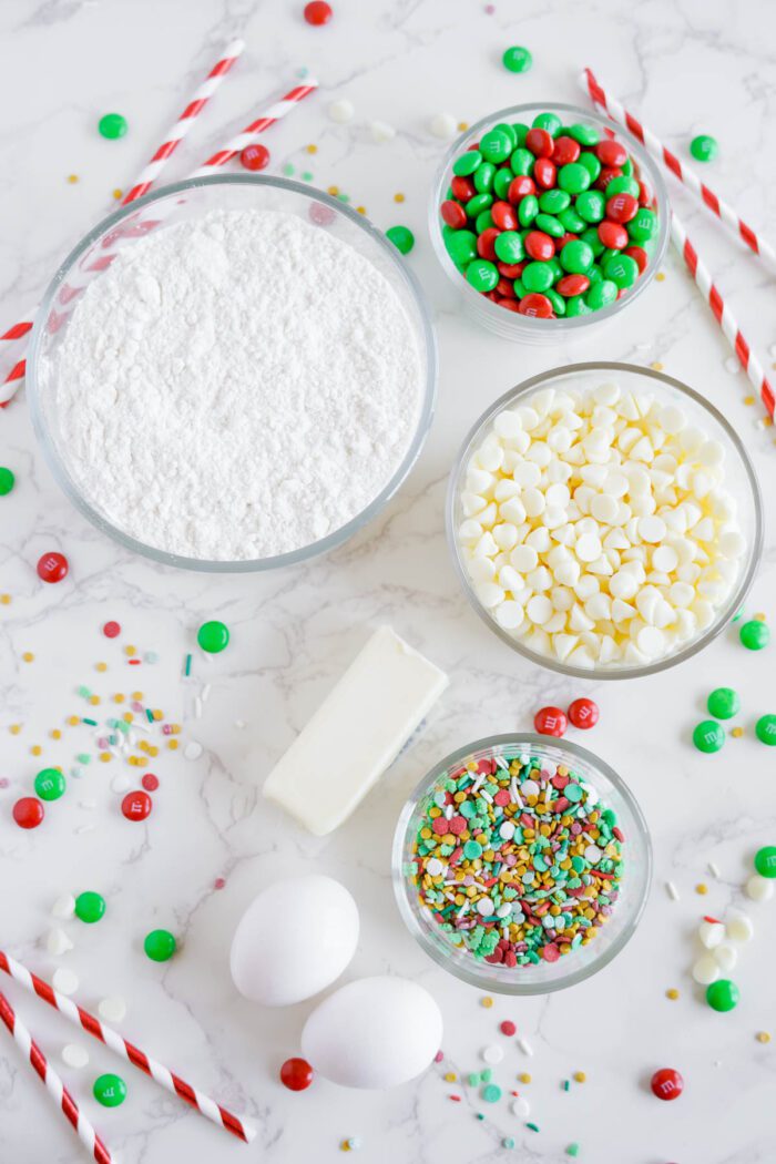 Candy cane straws, a bowl of powdered sugar, white chocolate chips, red and green M&M's, colorful sprinkles, a stick of butter, and two eggs arranged on a white marble surface.