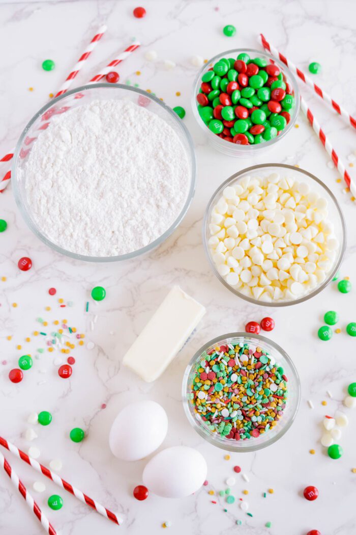 A baking setup with bowls of cake mix, green and red candies, white chocolate chips, sprinkles, two eggs, and a stick of butter on a white marble surface.