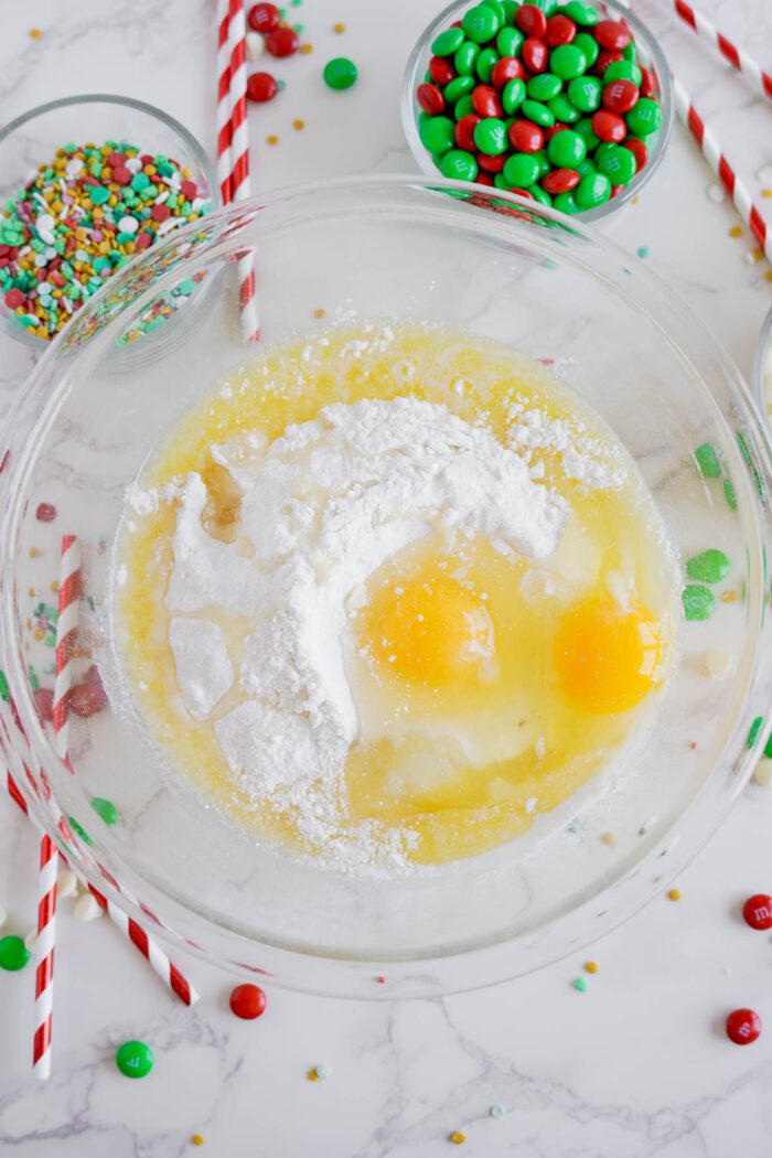 A mixing bowl containing ingredients like eggs, cake mix, and sugar, with bowls of colorful sprinkles and candy-coated chocolates and red-striped straws nearby on a marble countertop.