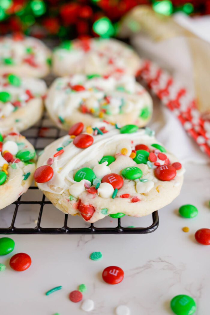 Christmas-themed cookies with white frosting and colorful candy pieces are displayed on a cooling rack. Red, green, and white candies are scattered around, with a festive background.