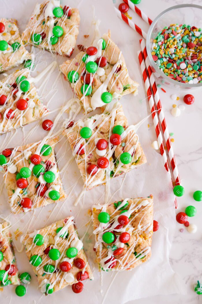 Overhead view of Christmas Cookie bars topped with green and red M&M candies and white drizzle. A small bowl of colorful sprinkles and two red-striped straws are on the side.