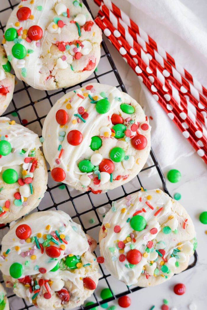 Cookies with white frosting and decorated with colorful candy pieces are displayed on a cooling rack, with red and white striped straws in the background.