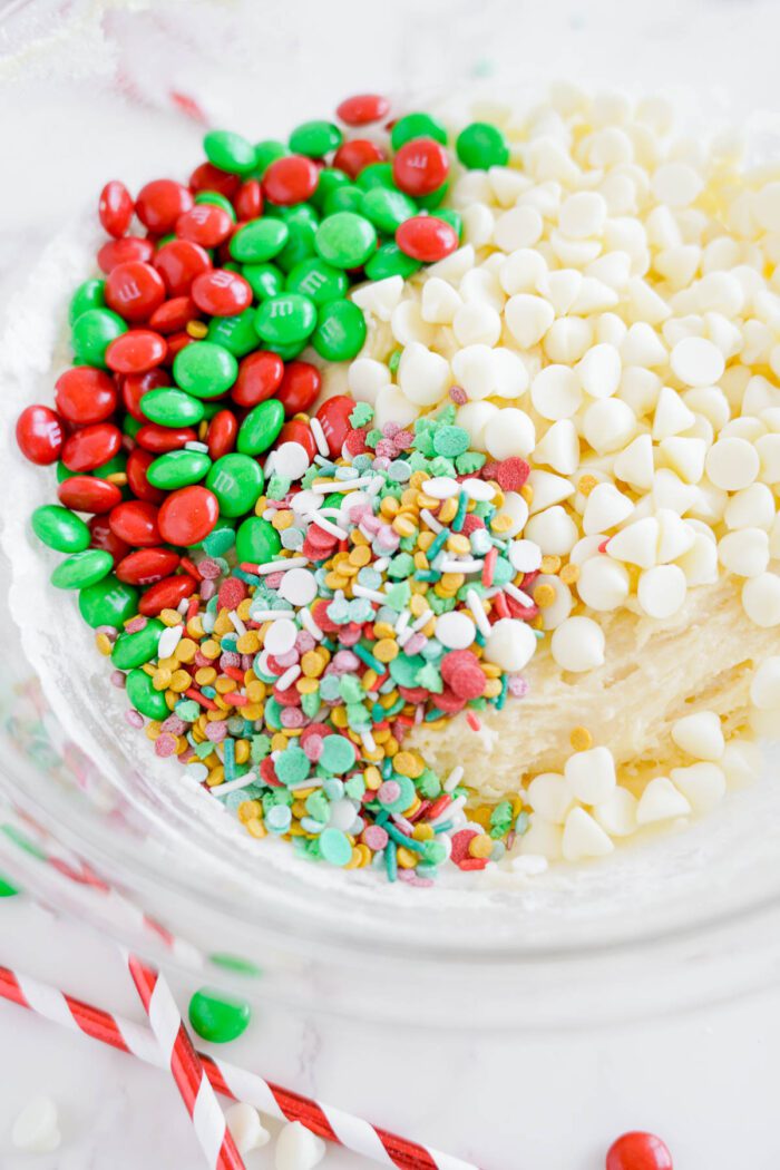 A glass bowl containing red and green M&Ms, white chocolate chips, and colorful sprinkles for baking. Two red and white striped candy canes are placed beside the bowl.