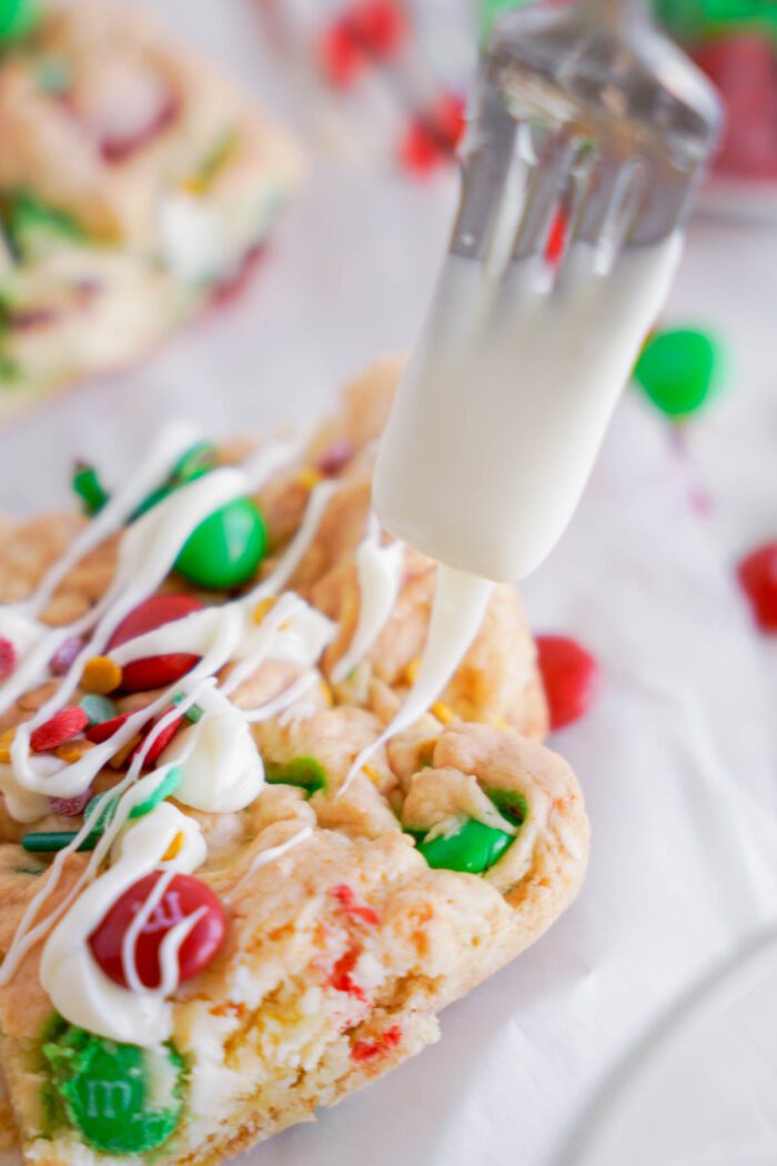 Close-up of a fork drizzling white icing onto a cookie topped with colorful candy pieces.