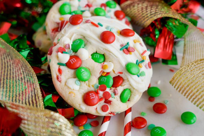 Christmas cookies with white frosting topped with red, green, and white M&Ms, surrounded by multicolored ribbon and a candy cane.