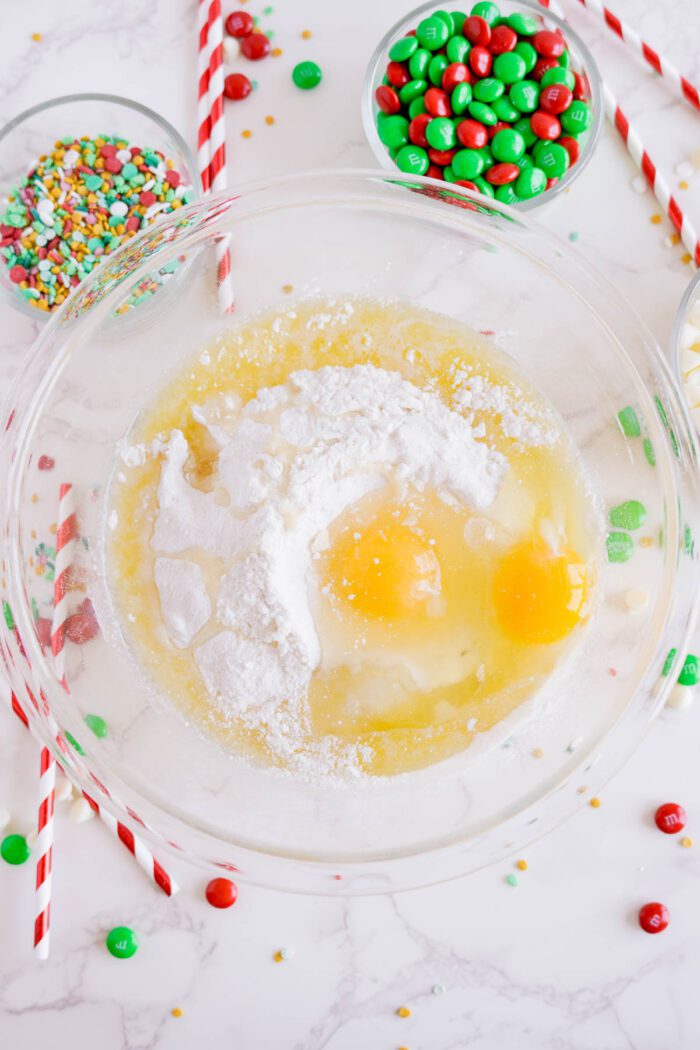 A glass bowl with cracked eggs, flour, and liquid ingredients is surrounded by bowls of colorful sprinkles, green and red candies, and candy-striped straws on a white marble surface.