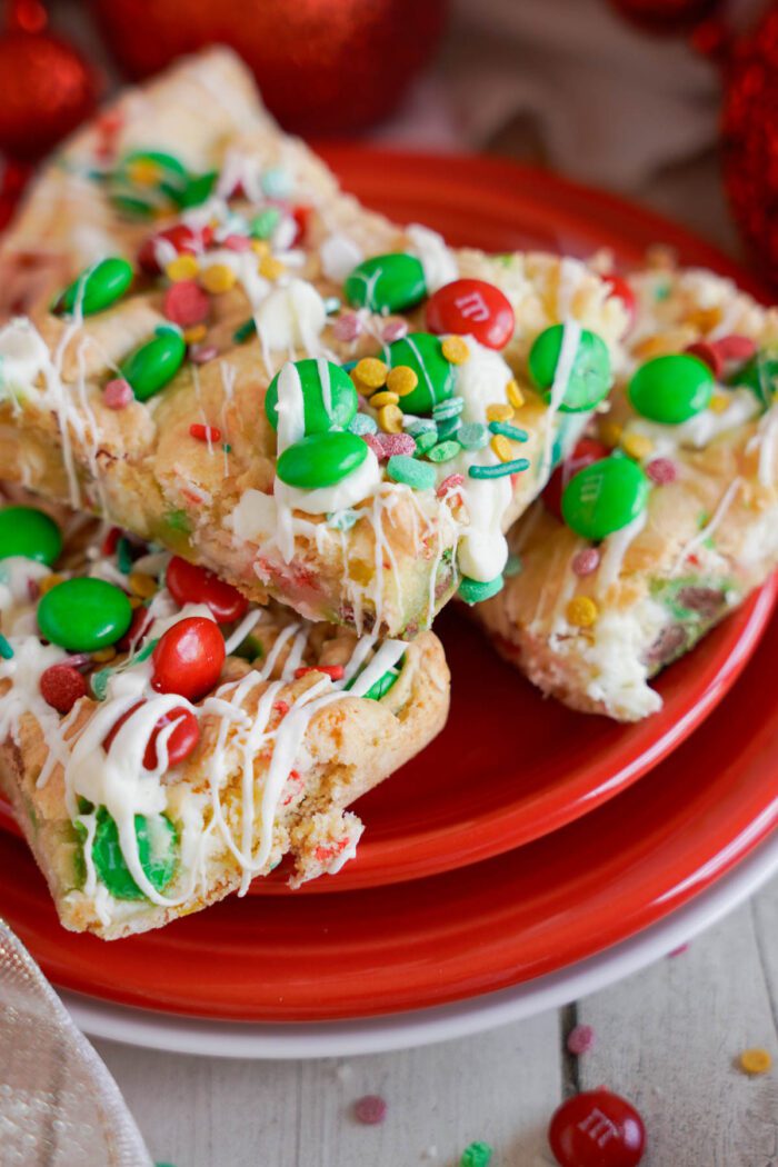 A red plate holds Christmas Cookie bars topped with green and red candy-coated chocolates, colorful sprinkles, and drizzled white icing.