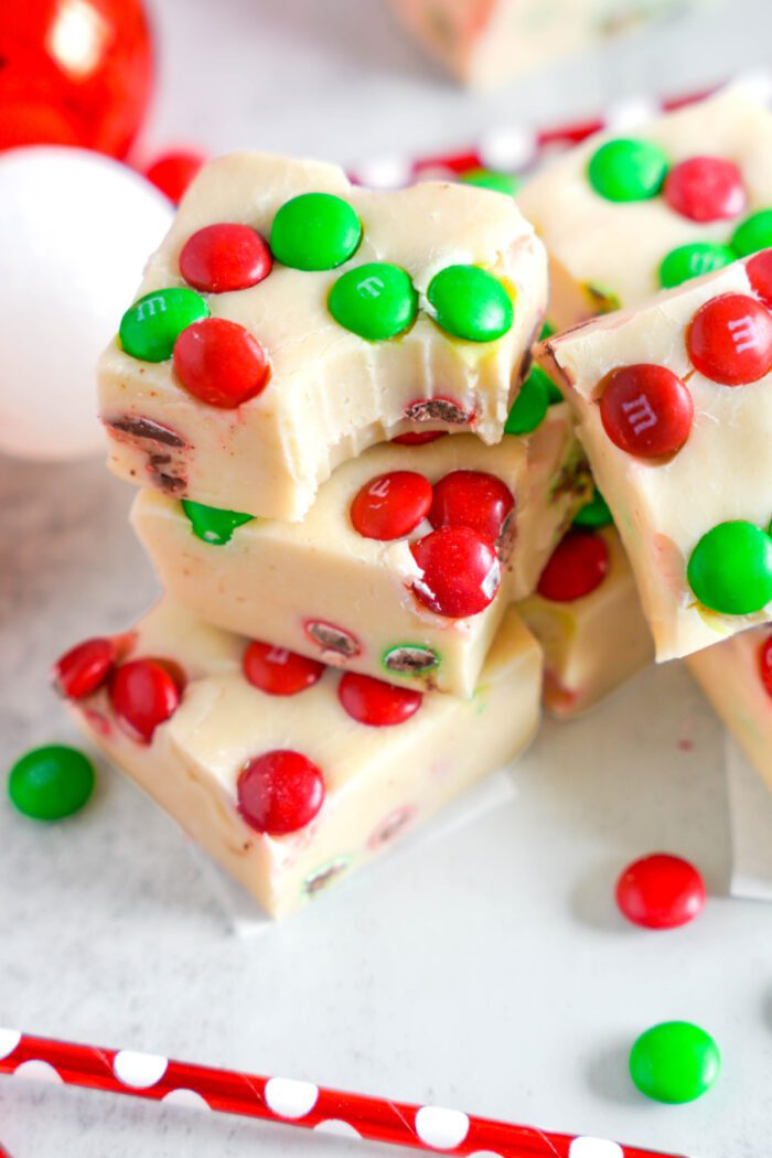 Close-up of white chocolate fudge squares with red and green M&M candies, stacked on a white surface with scattered M&Ms around.