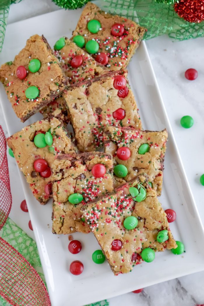 A white rectangular plate holds squares of holiday-themed cookie bars. The bars contain red and green M&M candies and festive sprinkles. Green and red ribbons are partially visible in the background.