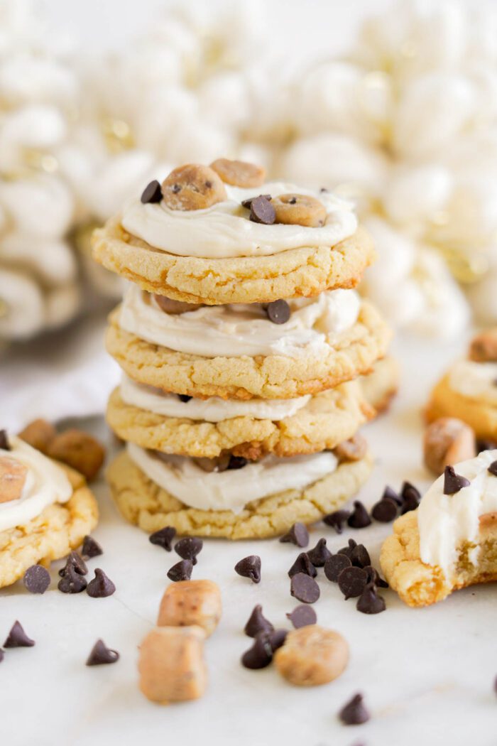 A stack of cookies with white frosting, topped with chunks of cookie dough and chocolate chips, surrounded by scattered chocolate chips and cookie pieces on a white surface—an easy cookie dough cookies recipe perfect for any occasion.