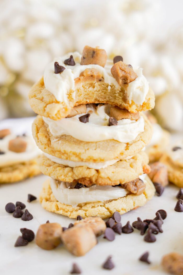 A stack of three frosted cookies with chunks of cookie dough and chocolate chips. The top cookie has a bite taken out of it. Additional cookie dough pieces and chocolate chips are scattered around, making this an easy Cookie Dough Cookies recipe that's both delicious and visually appealing.