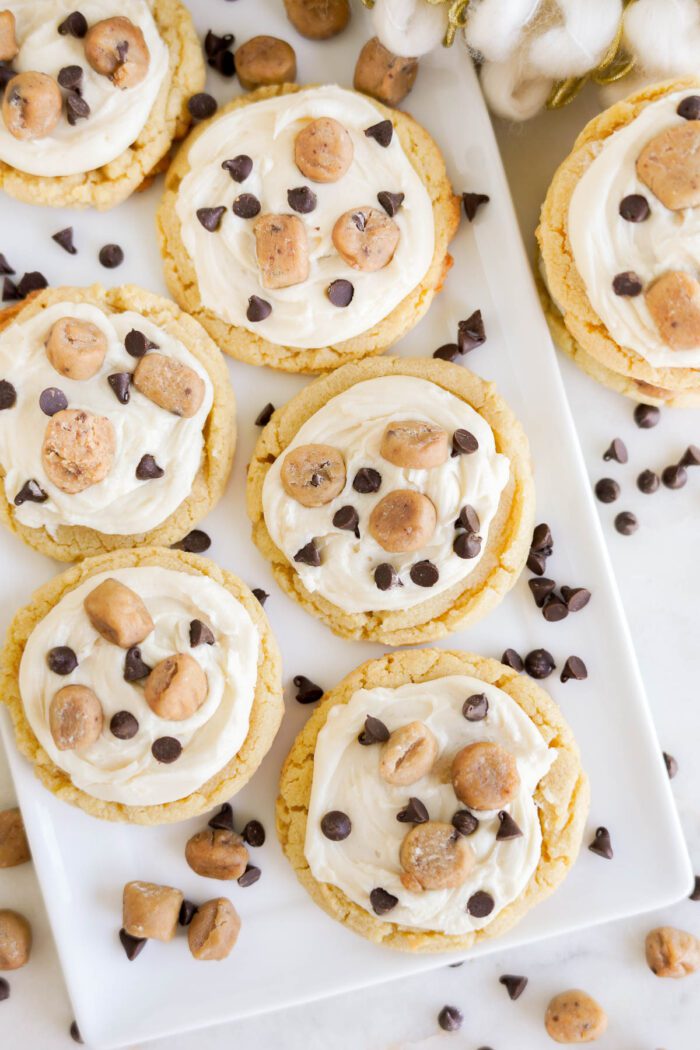 A white rectangular plate with seven cookies topped with white frosting, chocolate chips, and small cookie dough pieces, surrounded by scattered chocolate chips and cookie dough pieces from our Easy Cookie Dough Cookies Recipe.