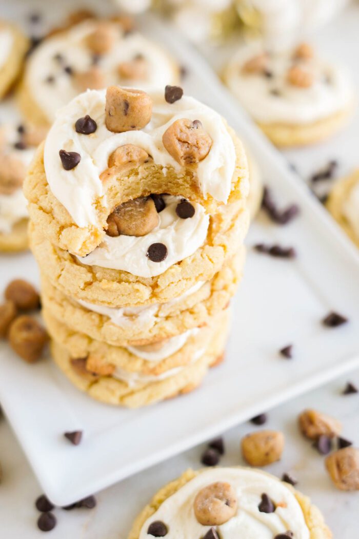 Stack of frosted cookies topped with cookie dough bites and chocolate chips, one cookie has a bite taken out of it, placed on a white plate with scattered chocolate chips around. Discover this easy Cookie Dough Cookies recipe for a delectable treat everyone will love.