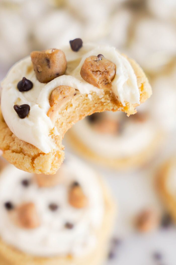 A bitten cookie with white frosting, chocolate chips, and cookie dough chunks is held up in focus—an irresistible treat straight from our Easy Cookie Dough Cookies Recipe.