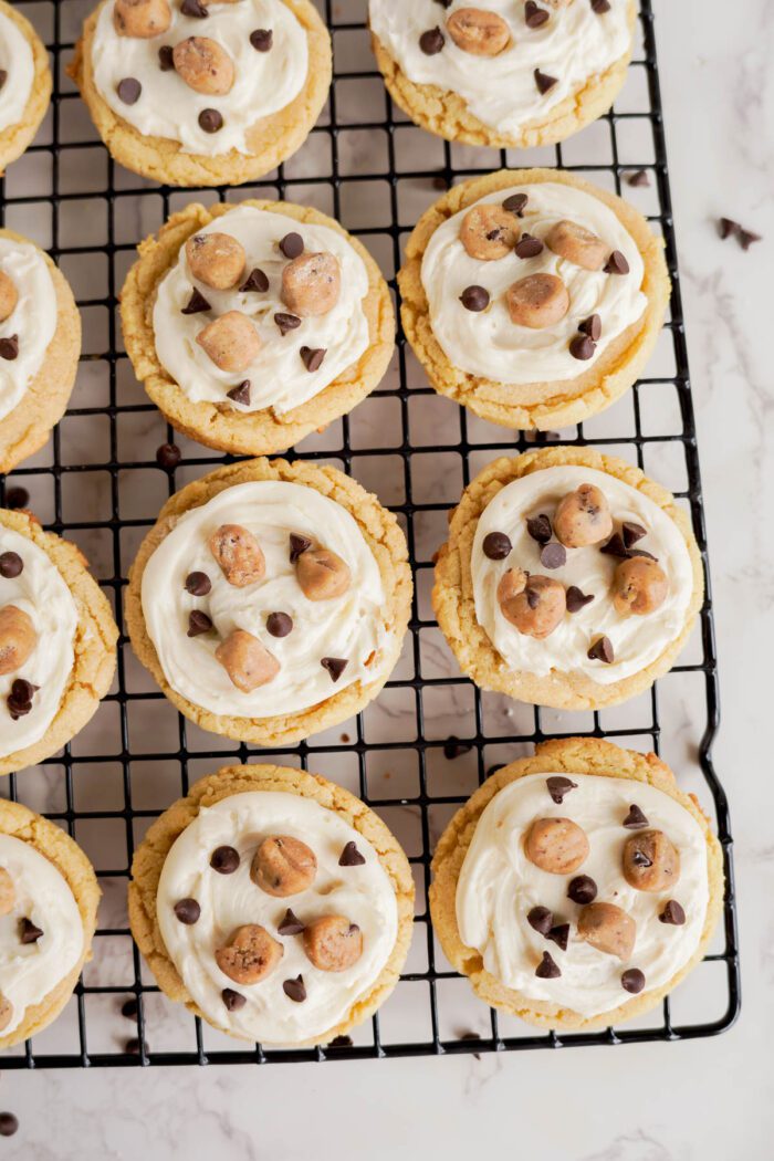 An easy cookie dough cookies recipe features twelve frosted cookies topped with cookie dough bites and chocolate chips, arranged on a cooling rack over a marble surface.