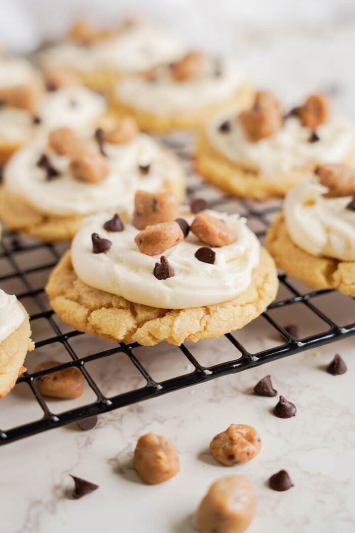 Frosted cookies topped with chocolate chips and small chunks of cookie dough are on a cooling rack. A few toppings are scattered on the surface below the rack, making this an Easy Cookie Dough Cookies Recipe perfect for any occasion.