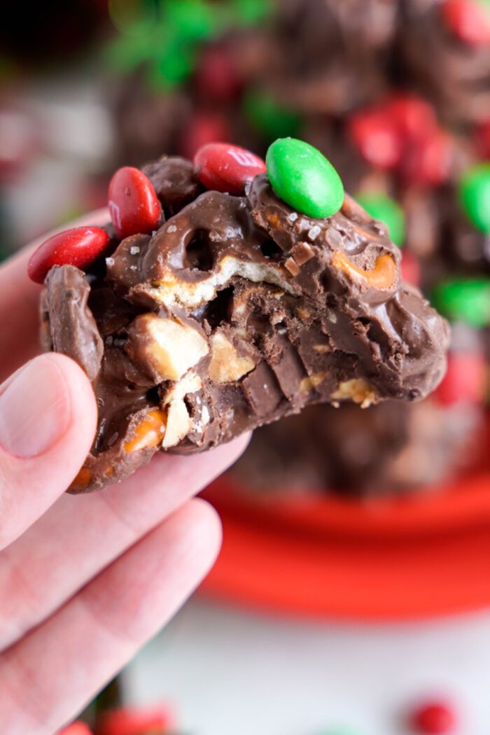 A hand holding a chocolate-covered treat with visible pretzel pieces and red and green candy-coated chocolates.