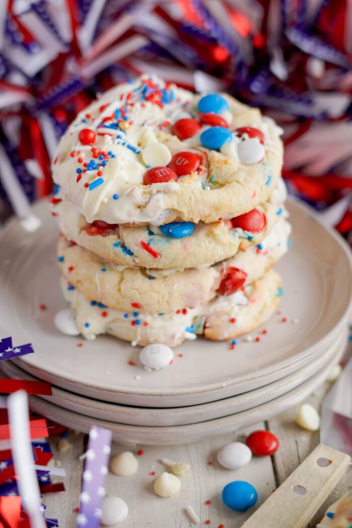 A stack of cookies decorated with white frosting, red, white, and blue sprinkles, and candy, placed on a white plate with patriotic ribbon decorations in the background.