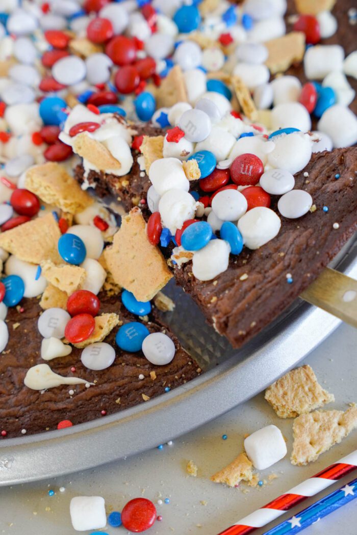 A close-up of a dessert pizza featuring a brownie base topped with red, white, and blue candies, mini marshmallows, and pieces of graham crackers on a metal pizza pan.