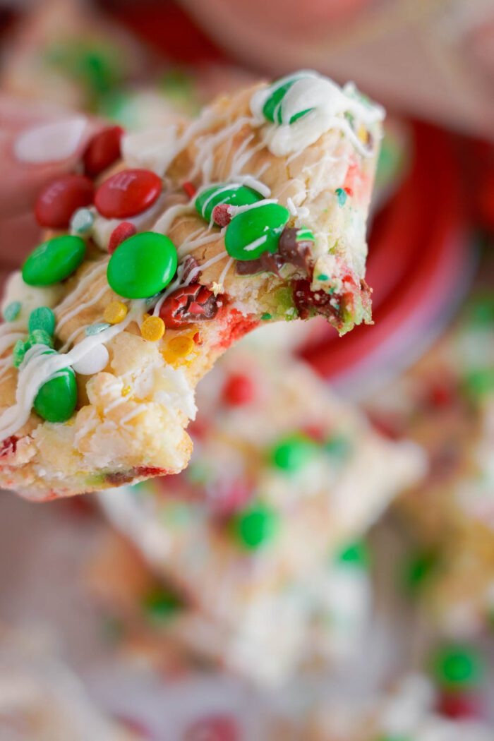 A close-up of a Christmas Cookie Bar treat topped with colorful red, green, and white candy pieces, drizzled with white icing. The treat has a bite taken out of it.