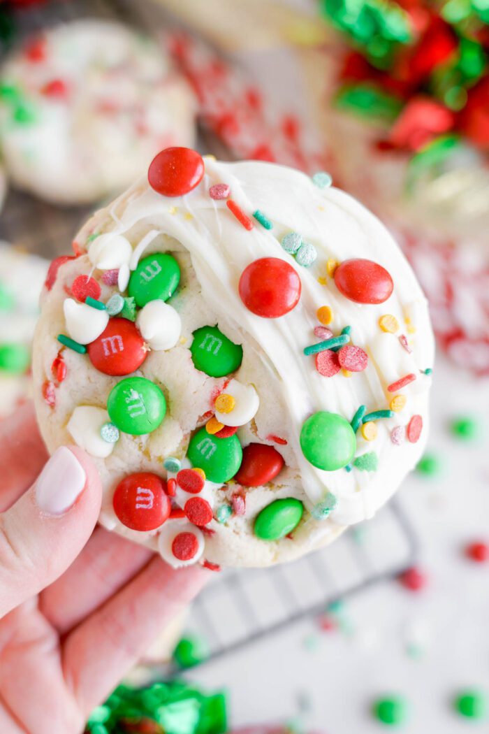 A hand holding a festive cookie topped with white frosting, red and green M&M's, and colorful sprinkles.