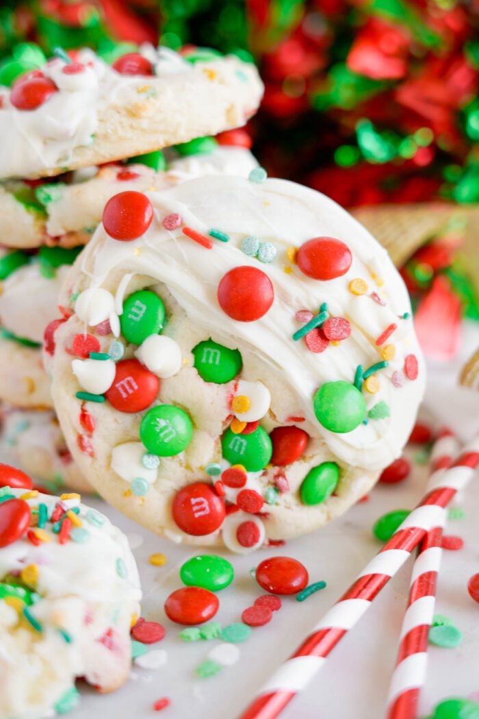 Colorful holiday cookies topped with white chocolate and a variety of red, green, and white M&Ms and sprinkles, with festive, blurred holiday decorations in the background.