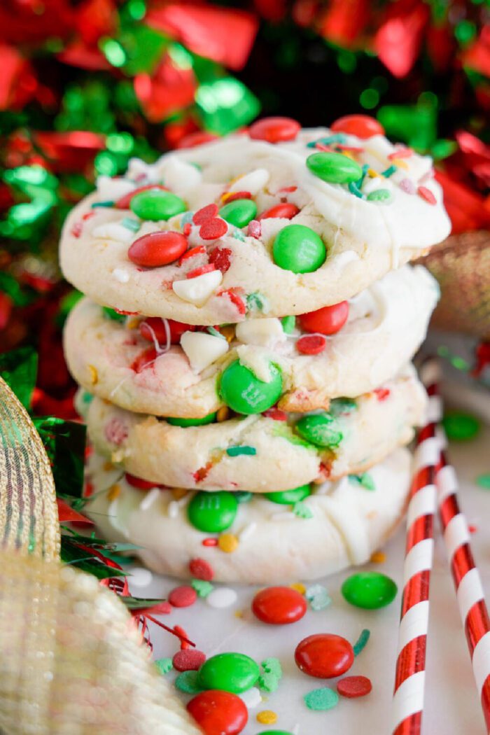 A stack of five colorful Christmas cookies with green and red candies and sprinkles, surrounded by festive red and green decorations.