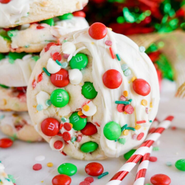 A close-up of a festive cookie decorated with red and green M&Ms, white chocolate, and colorful sprinkles, against a backdrop of holiday decorations.