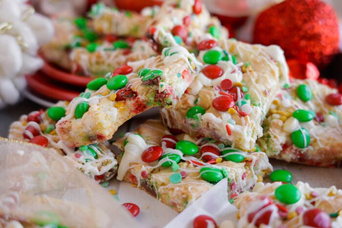 A pile of Christmas Cookie bars topped with red and green candy-coated chocolates and white chocolate drizzle on a table.