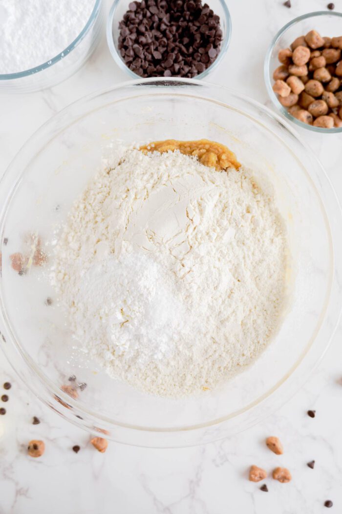 A mixing bowl with flour and other ingredients sits on a marble surface, surrounded by small bowls containing chocolate chips and nuts, perfect for whipping up an easy cookie dough cookies recipe.