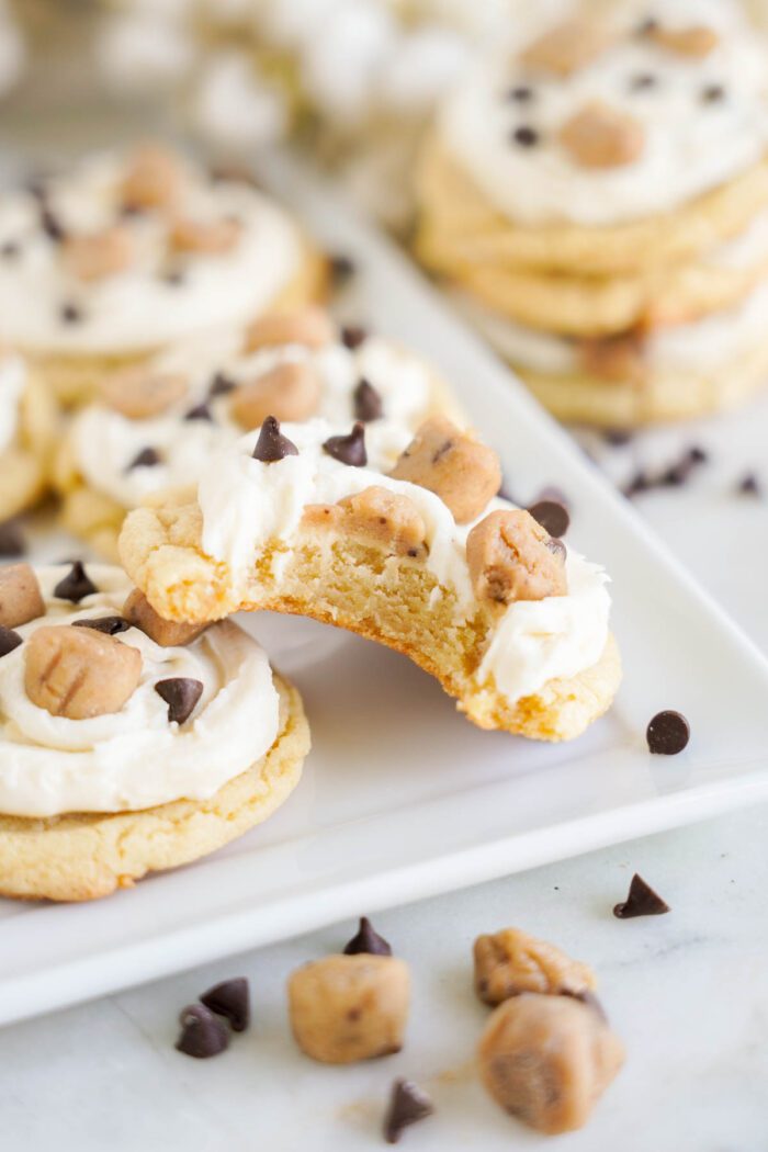 A plate of cookies topped with frosting, chocolate chips, and cookie dough chunks from an easy cookie dough cookies recipe. One cookie in the foreground has a bite taken out of it.