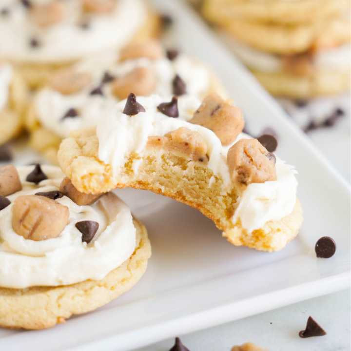 A frosted cookie topped with cookie dough chunks and chocolate chips has a bite taken out of it. Other cookies are in the background on a white rectangular plate, demonstrating the perfection of this Easy Cookie Dough Cookies recipe.