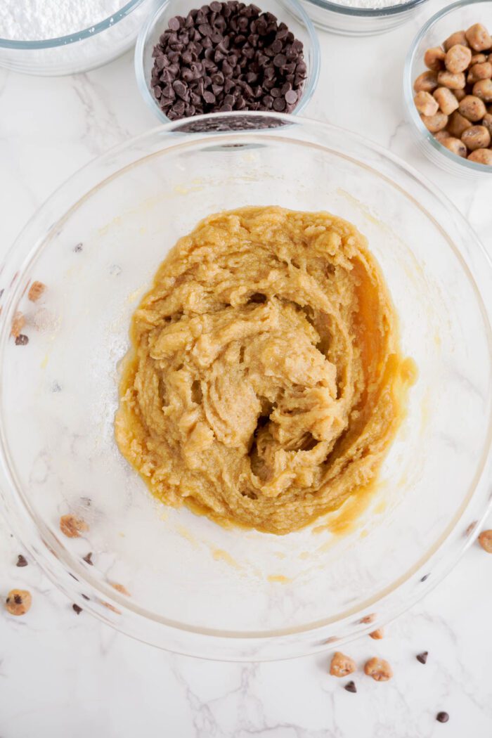 A glass mixing bowl filled with light brown cookie dough, surrounded by bowls containing chocolate chips, hazelnuts, and powdered sugar on a marble surface—an essential setup for an easy cookie dough cookies recipe.