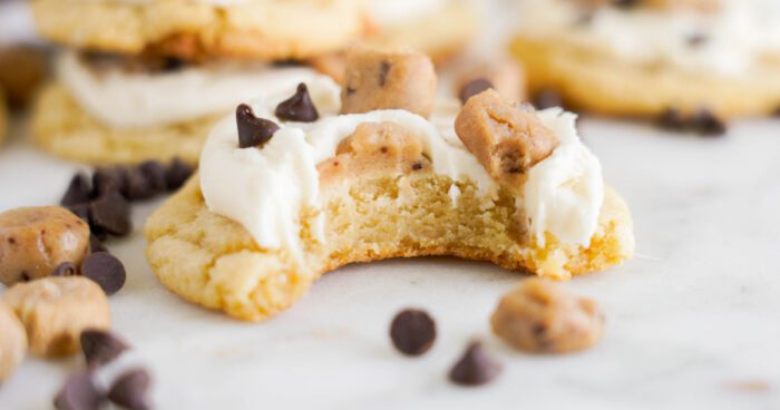 A close-up of a partially eaten cookie topped with white frosting, chocolate chips, and chunks of cookie dough. Chocolate chips are scattered around on the surface, embodying an easy Cookie Dough Cookies recipe.