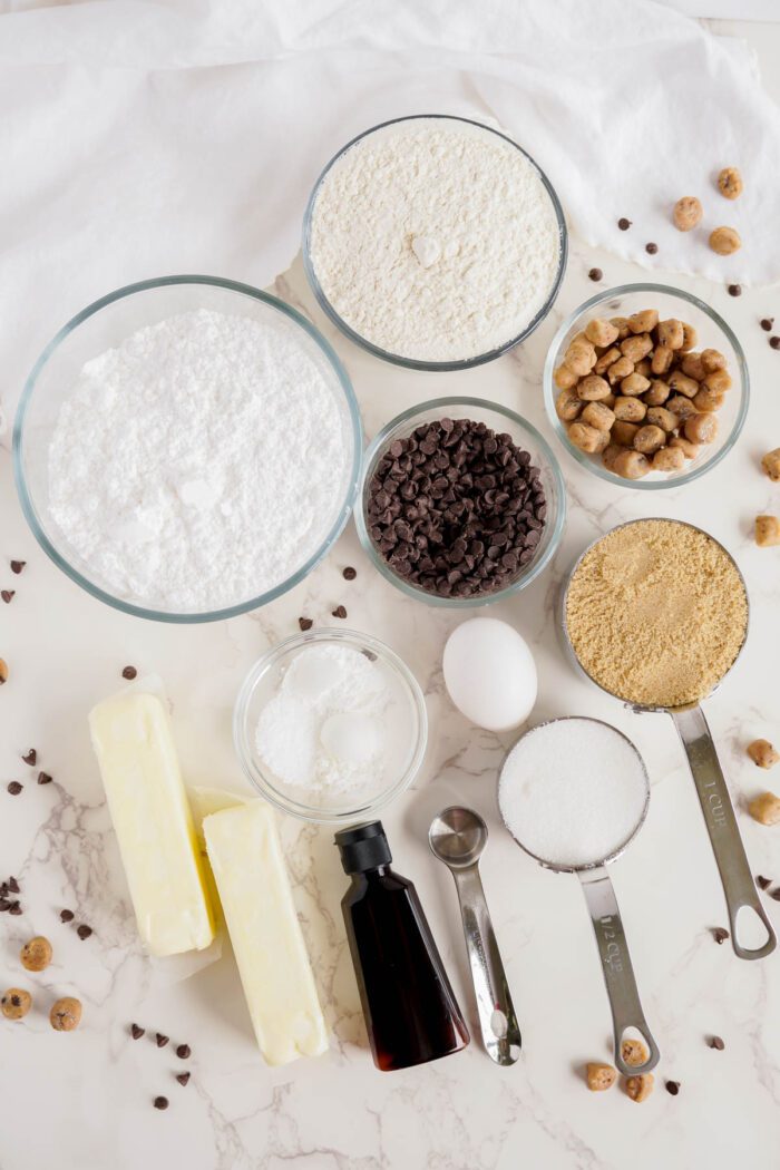 A top-down view of various baking ingredients, including flour, sugar, butter, chocolate chips, an egg, baking soda, vanilla extract, and nuts arranged in bowls and measuring tools on a marble surface—everything you need for an easy cookie dough cookies recipe.