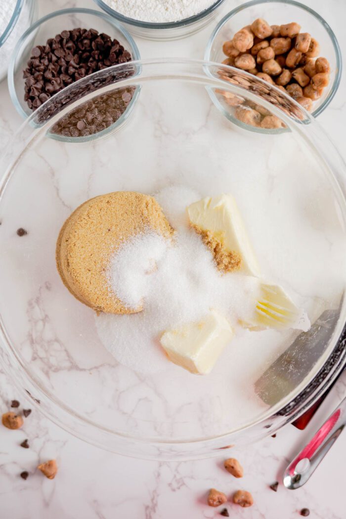 A clear glass bowl containing brown sugar, white sugar, and butter sits on a white marble surface. Surrounding it are smaller bowls with chocolate chips, flour, and nuts—ideal ingredients for an easy cookie dough cookies recipe. A red utensil rests nearby.