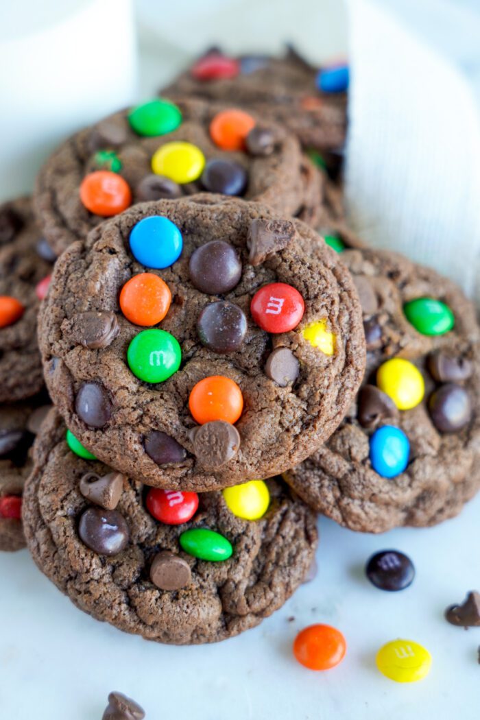 A pile of chocolate cookies topped with colorful candy-coated chocolates and chocolate chips on a white surface.