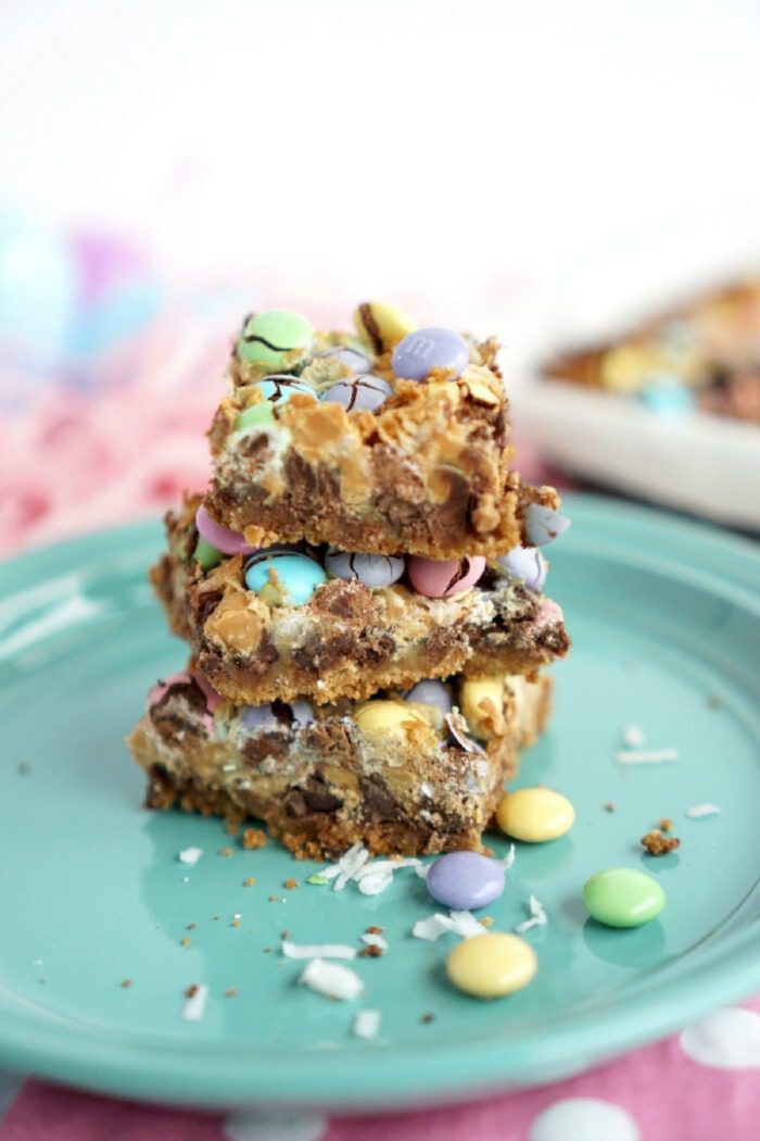 Three stacked dessert bars with colorful candy-coated chocolates, chocolate chips, and a toasted topping sit on a green plate, with additional candy pieces scattered around.