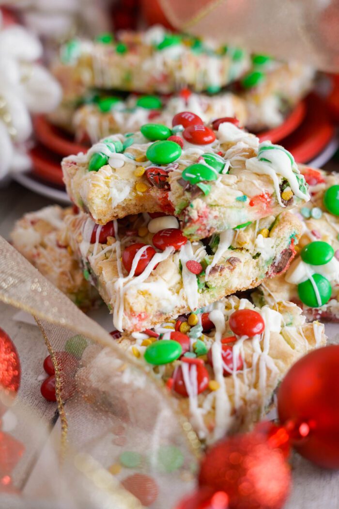 A close-up of colorful Christmas Cookie bars topped with white drizzle, red and green candies, and festive sprinkles, surrounded by red ornaments and ribbon.