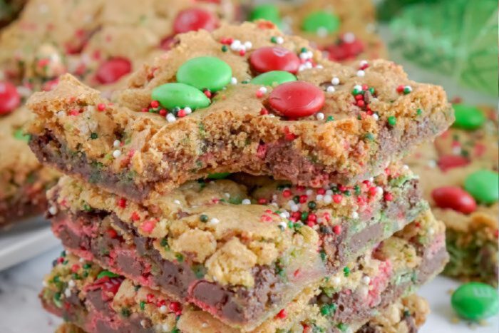 A stack of cookie bars topped with red and green candy-coated chocolates and sprinkles. The cookie bars have a chocolate layer and appear to have a festive holiday theme.