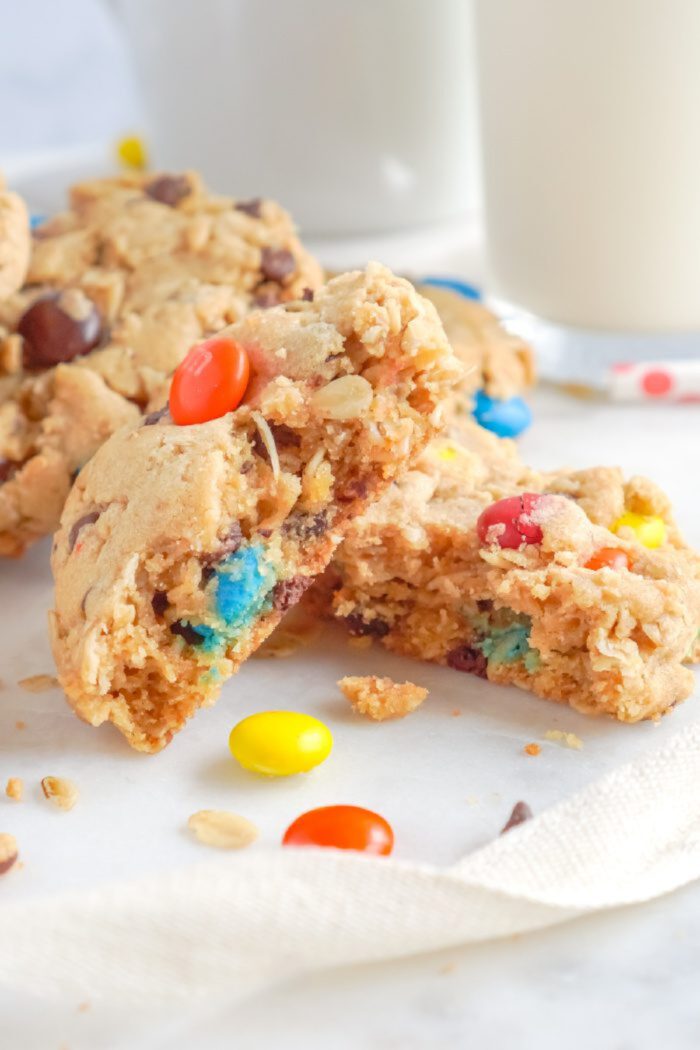 Cookies with colorful candy pieces are placed on a white surface. Some cookies are broken, revealing their interior. A glass of milk is in the background.