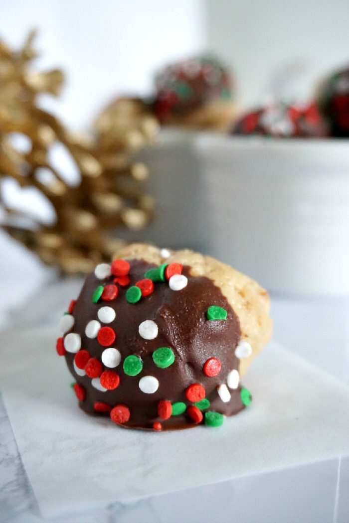 A holiday-themed peanut butter ball covered in chocolate and decorated with red, green, and white sprinkles sits on a piece of parchment paper next to a bowl of similar treats, perfect for featuring in peanut butter dessert recipes.