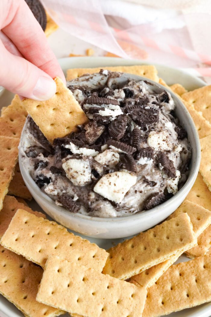 A bowl of Oreo dip surrounded by golden crackers, with a hand dipping a cracker into the dessert.