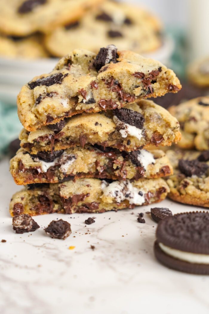 A stack of cookies with chunks of chocolate and crushed oreo pieces, set against a blurred background of more cookies.