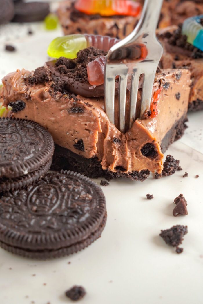 A slice of Oreo chocolate cheesecake topped with gummy candies and crushed cookies, with a fork digging in, on a white plate.